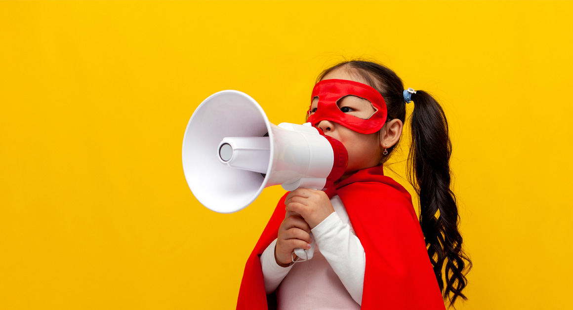 little Asian girl in superman costume and mask announces news and information into loudspeaker, Korean child in superhero cape shouts to the side on yellow isolated background