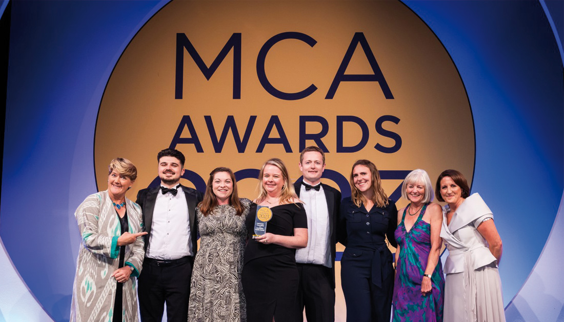 A photograph of 8 people in front of the "MCA Awards 2023" backdrop. One of the group is holding the MCA award for Performance Improvement in the Public Sector.