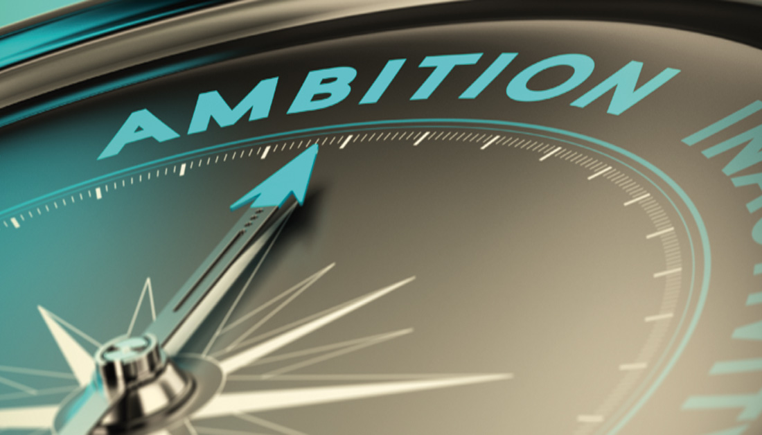 Close-up of a compass dial with the word “AMBITION” prominently displayed across the top in teal lettering. The compass needle points decisively forward, suggesting direction, drive and purpose. The metallic finish and soft lighting create a polished, aspirational feel, reinforcing themes of leadership, strategy and forward momentum.