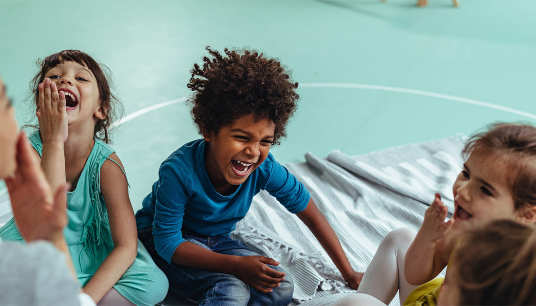 Image of a group of young children sitting together on a gym floor, laughing and clapping during a playful activity. The setting features a soft green indoor sports surface and a light blanket spread beneath them. The children are smiling broadly and interacting with someone just out of frame, capturing a joyful, energetic moment of shared play and social connection.