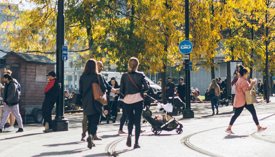 A lively urban street scene on a sunny day. People are walking along tram tracks lined with tall trees covered in bright yellow autumn leaves. Several pedestrians are crossing the street, including a woman pushing a pram. Others stand chatting or sitting in the background near small kiosks and buildings. The atmosphere feels relaxed and social, with warm sunlight and a sense of everyday city life.