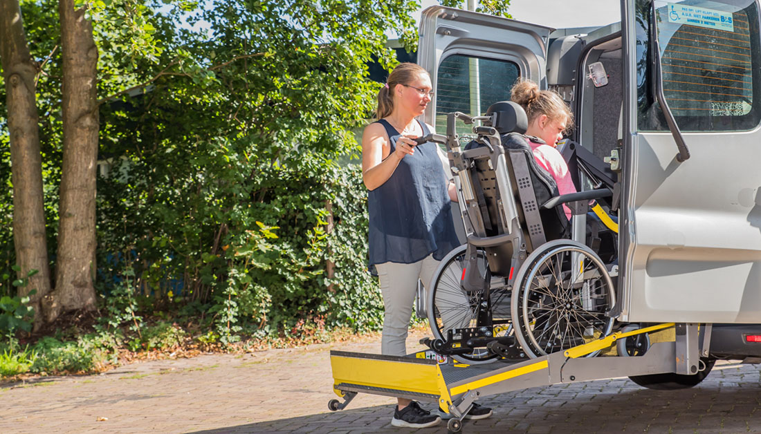 Image of a woman assisting a wheelchair user to board an accessible minibus using a rear hydraulic lift. The scene is outdoors on a sunny day, with trees and greenery in the background. The wheelchair is positioned securely on the yellow platform lift as it is raised into the vehicle, highlighting inclusive transport, mobility support and independent travel.