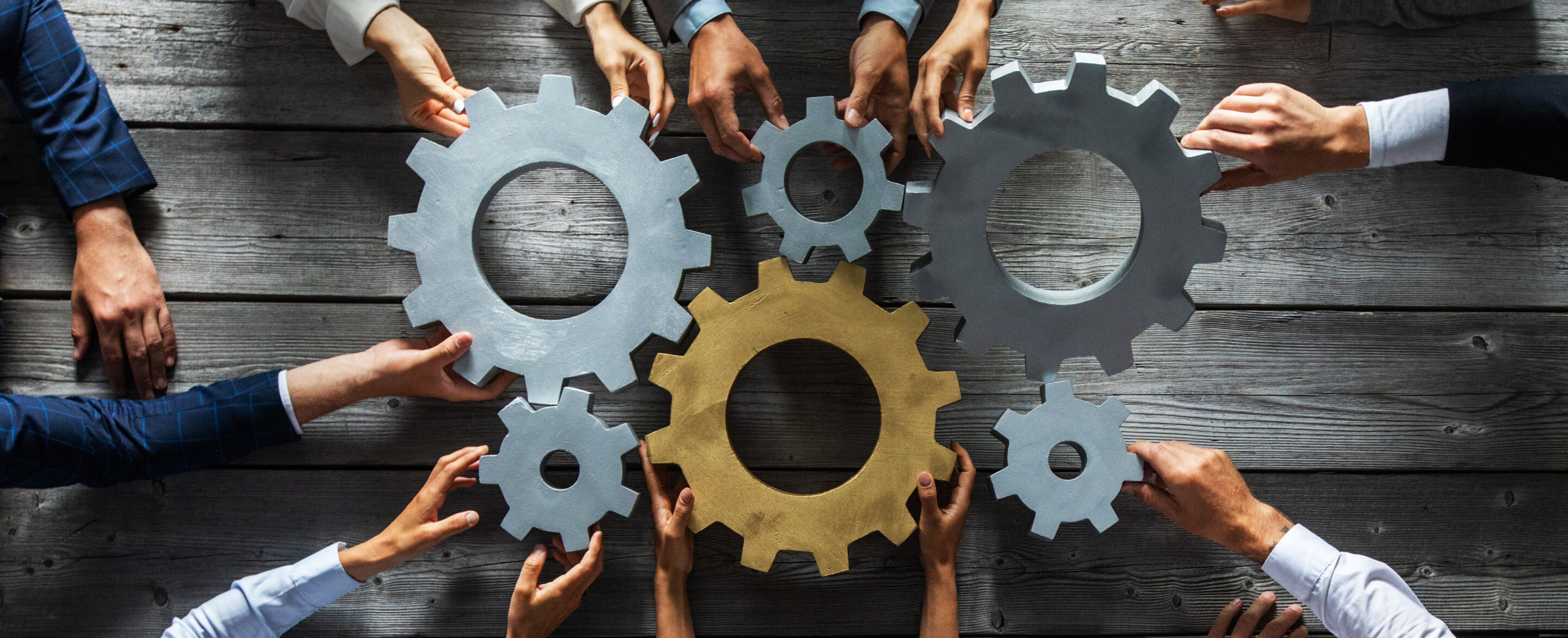 Multiple people reach in from different sides to hold large interlocking gear wheels on a wooden surface, symbolising teamwork, collaboration, and coordinated effort.