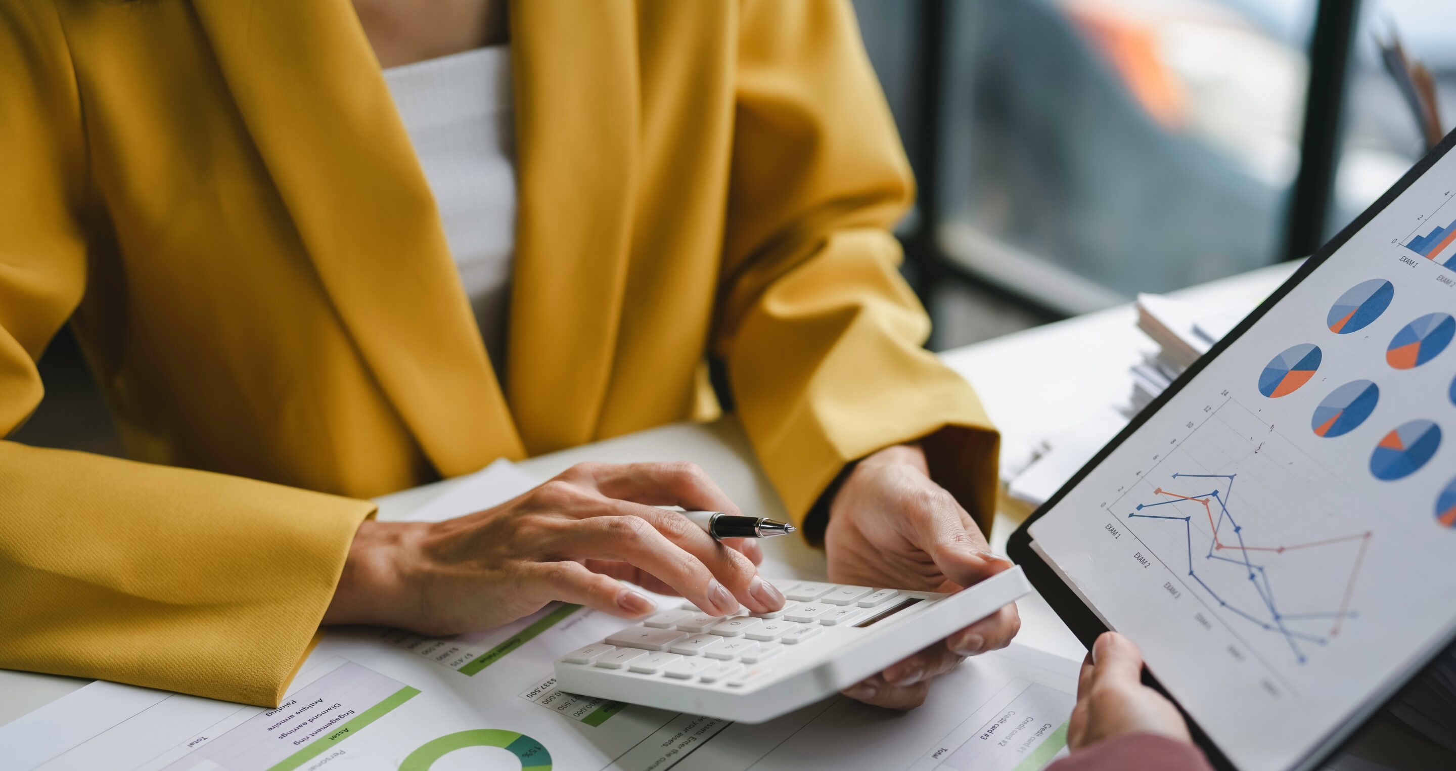 A person in a yellow blazer uses a calculator while reviewing printed financial charts, as another person holds a tablet displaying colourful graphs and data.