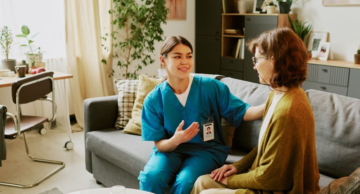 A healthcare professional in scrubs sits on a sofa beside a person, speaking with them and offering support in a home-like living room setting. The professional has one hand on the person's shoulder in a reassuring manner.