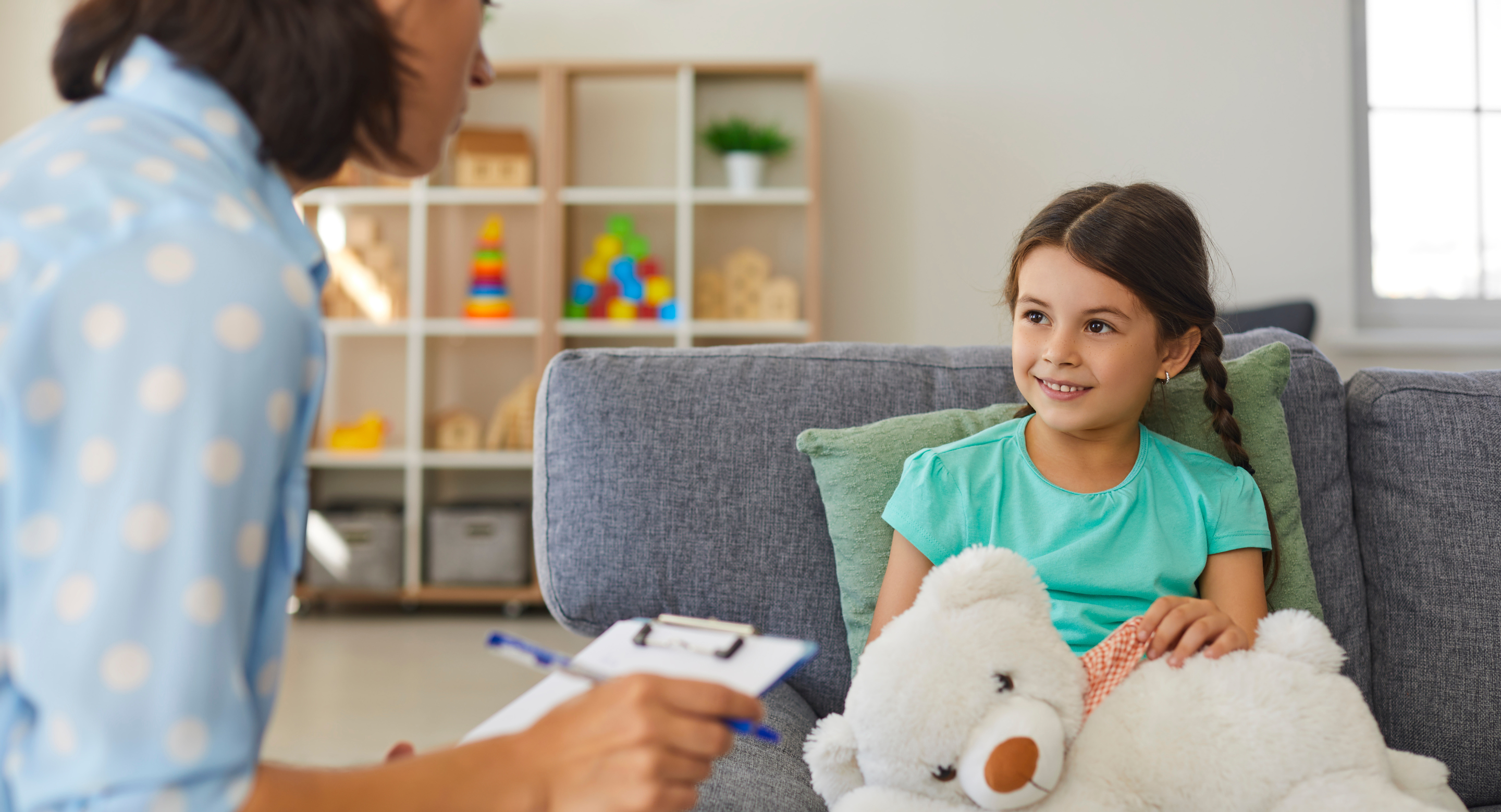 A young girl with braided hair sits on a grey couch holding a white teddy bear, smiling during a session with a female professional who is holding a clipboard. The background shows shelves with colourful toys, suggesting a child-friendly therapy or counselling setting.