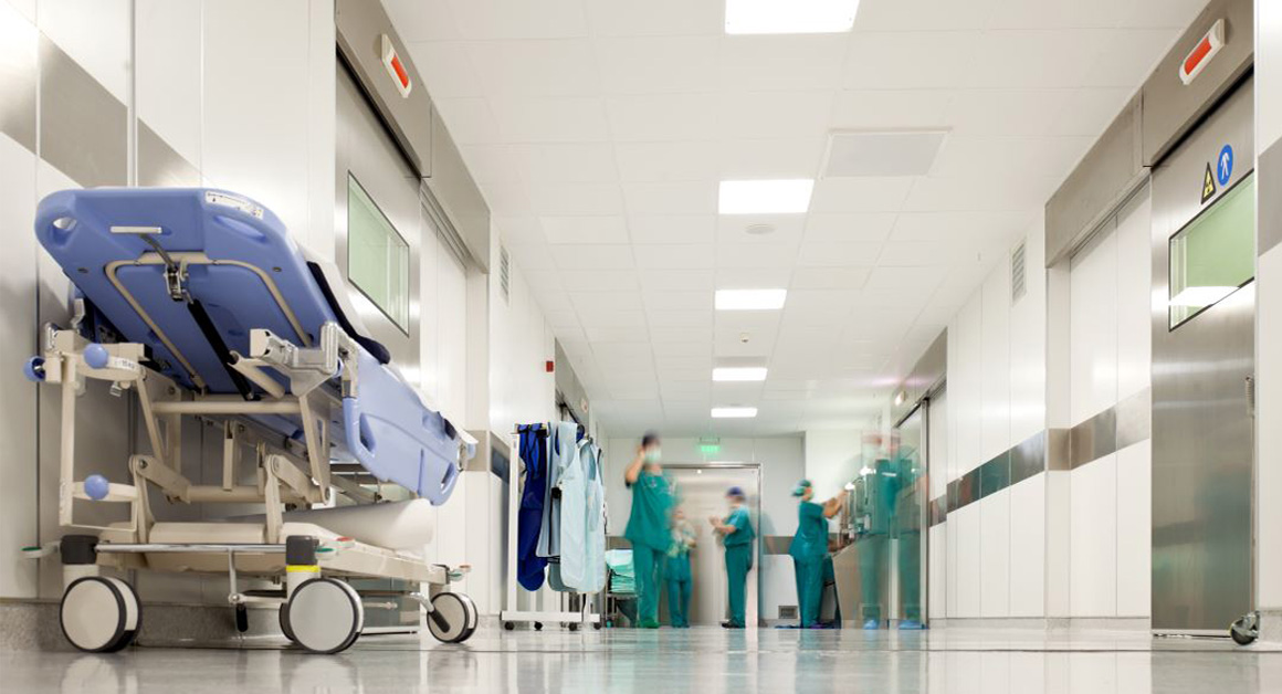 A bright hospital corridor with a wheeled hospital bed in the foreground, while several healthcare staff in green scrubs stand and move in the background near doors and equipment.
