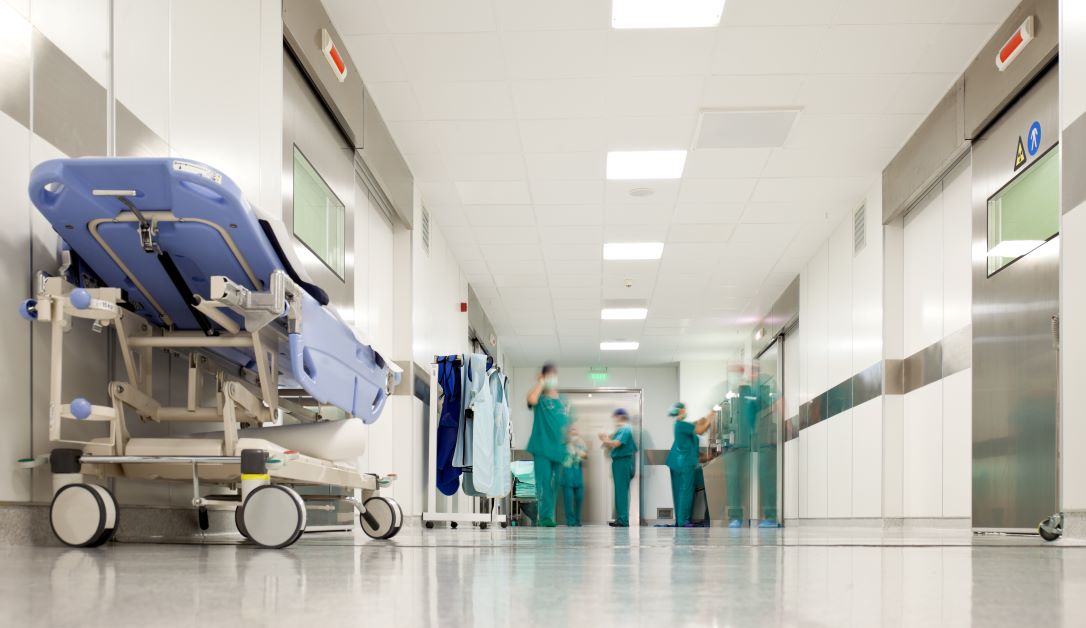 Hospital corridor with a patient stretcher on the left and medical staff in scrubs walking and talking in the distance, conveying a busy clinical environment.