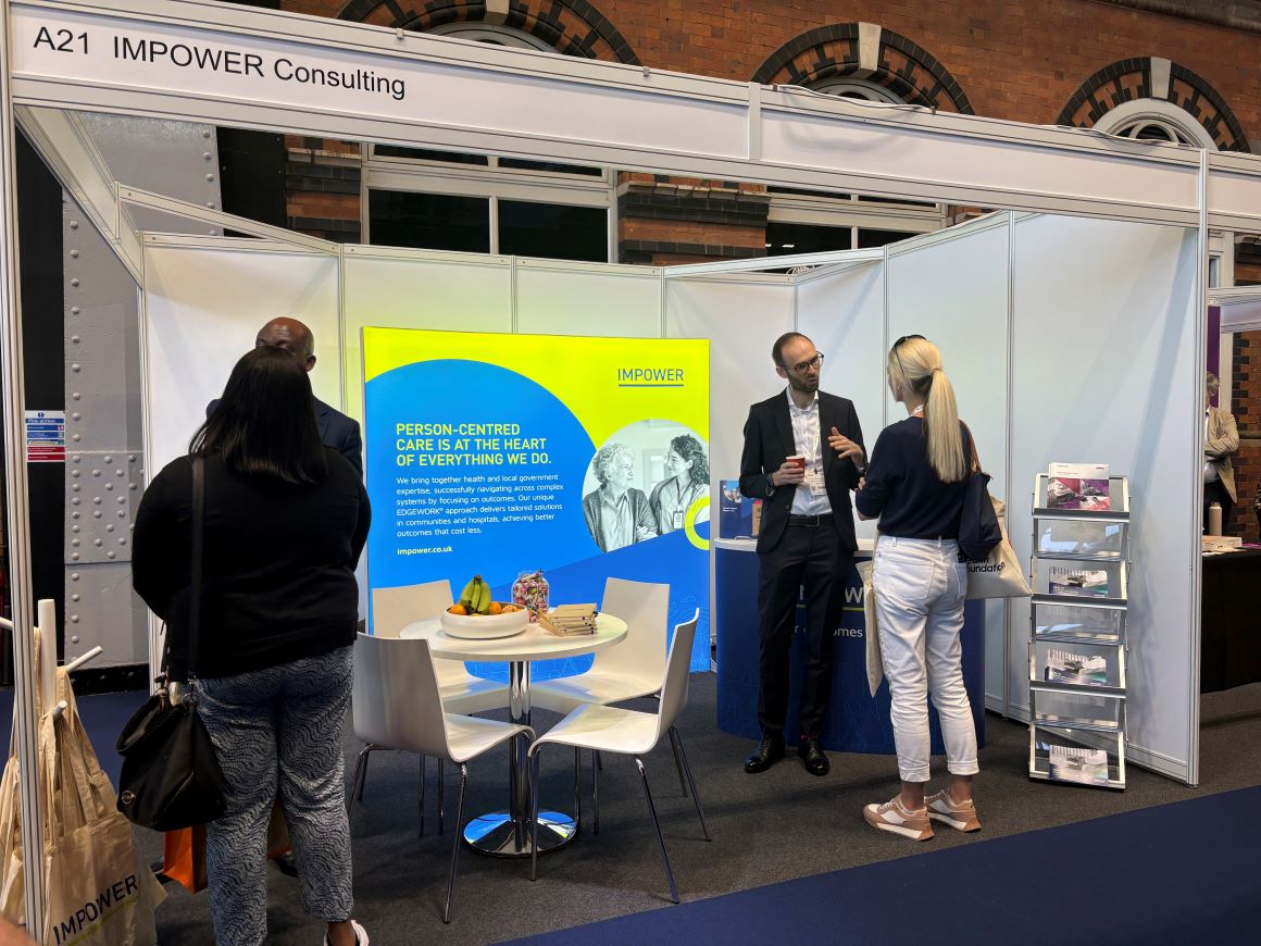 Four people speaking to each other on the IMPOWER Consulting exhibition stand at NHS ConfedExpo 2025, featuring a bright blue and lime green display that reads “Person‑centred care is at the heart of everything we do,” with a small table and chairs in front and brochures displayed to the right.