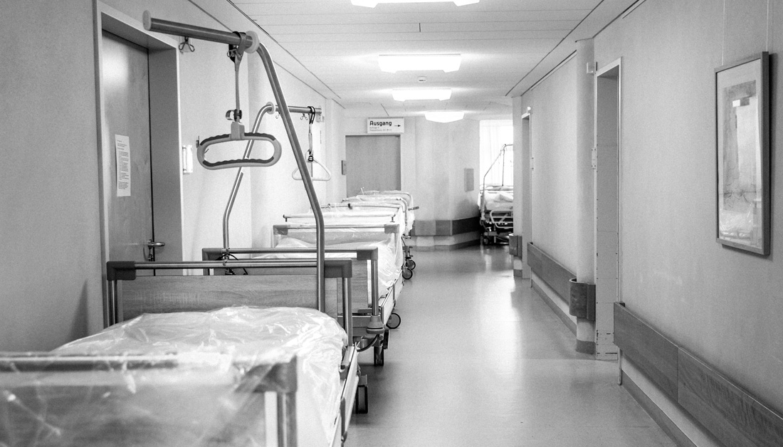 Black and white photo of a hospital hallway lined with empty hospital beds covered in plastic sheets, some with patient lifting handles attached.