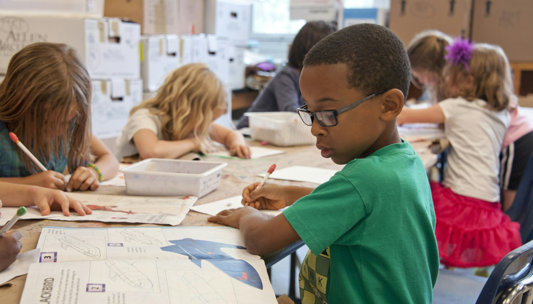Group of young children sitting at a classroom table working on drawings and assignments, with papers and pencils spread out; a boy in a green shirt and glasses focuses on his work in the foreground