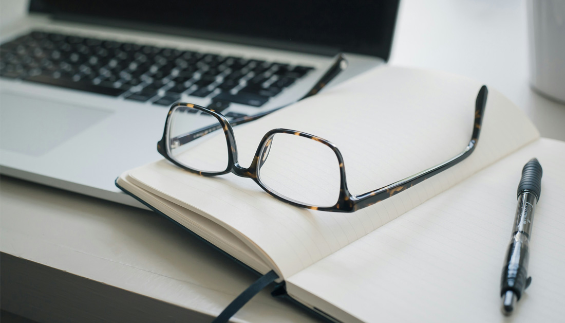 A pair of glasses resting on an open notebook beside a laptop, with a pen placed on the page. The scene suggests focused work, note-taking, or study in a clean, modern workspace.