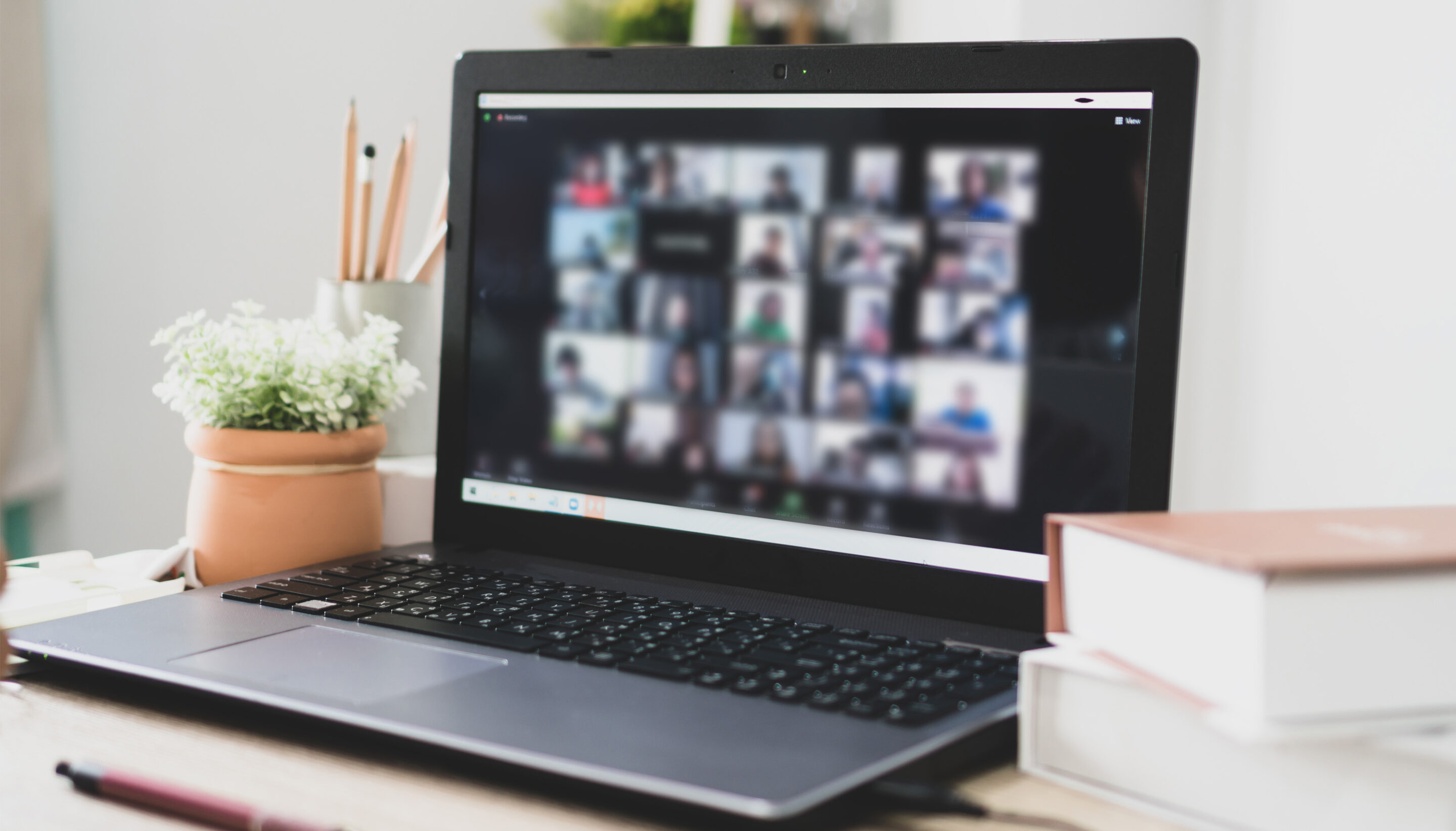 A laptop on a desk displaying a video call with multiple participants shown in a grid on the screen. The workspace includes a small potted plant, pencils in a holder, and stacked books, suggesting a remote meeting or online collaboration in a home or office setting.
