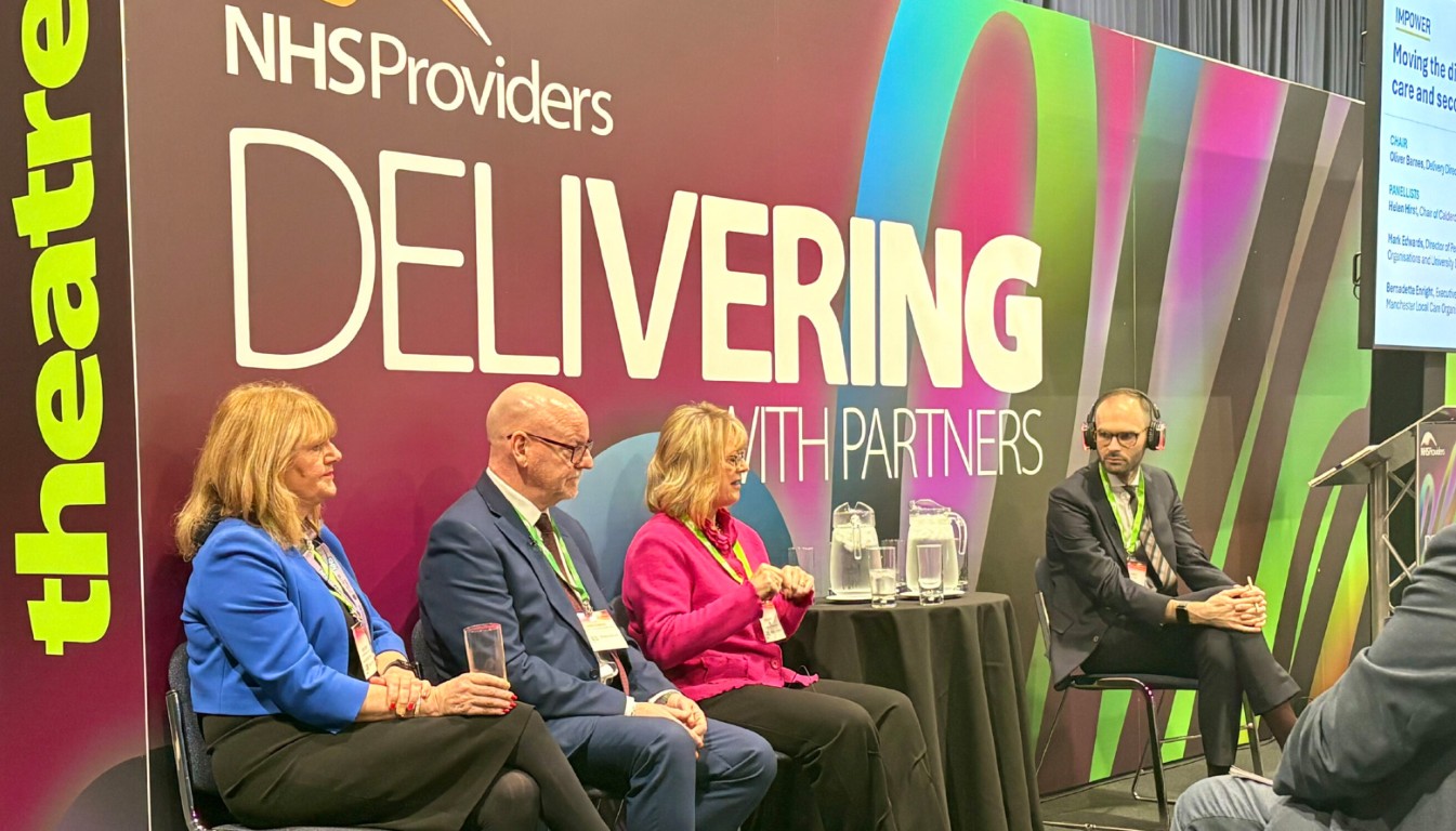 Panel discussion at an NHS Providers conference, with four speakers seated on stage in front of a large, colourful backdrop reading “Delivering with partners.” The panelists are wearing conference badges and speaking into microphones, with a small table holding water jugs between them, creating a professional event setting.
