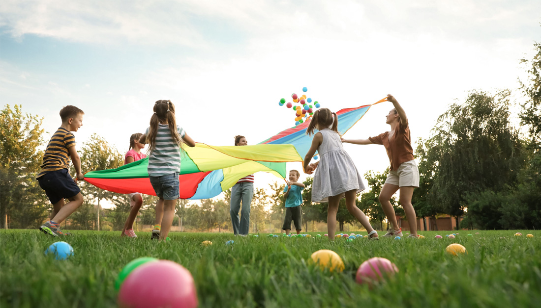 A group of children playing together outdoors on a grassy field, lifting and running with a large, colourful parachute. Bright balls are scattered on the grass, and the scene feels energetic, joyful, and inclusive under a clear sky.