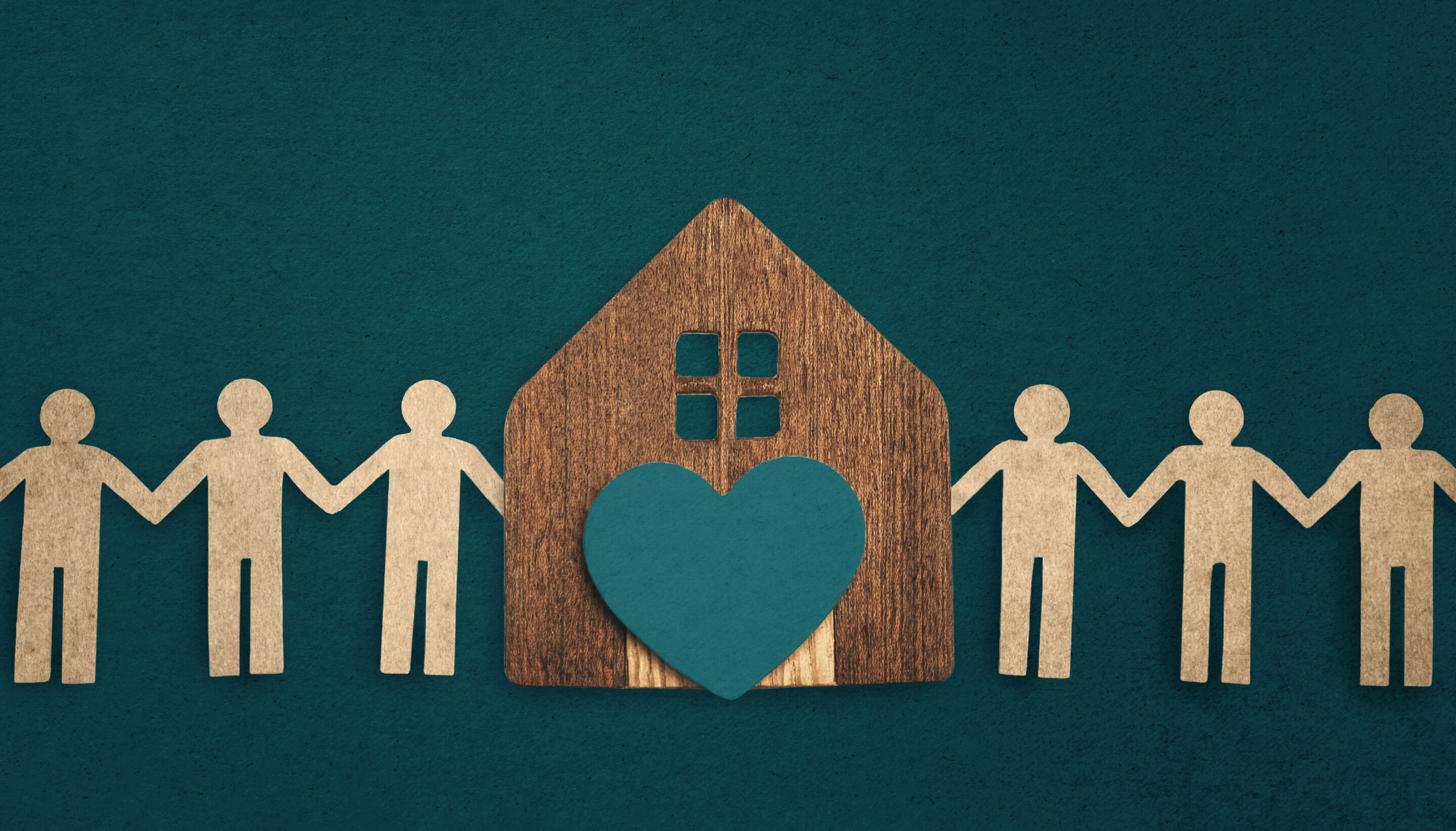 An illustration showing a row of simple paper-cut figures holding hands on either side of a wooden house. A heart shape is positioned on the front of the house, symbolising care and wellbeing. The background is a deep teal colour, giving the image a calm, community-focused feel.