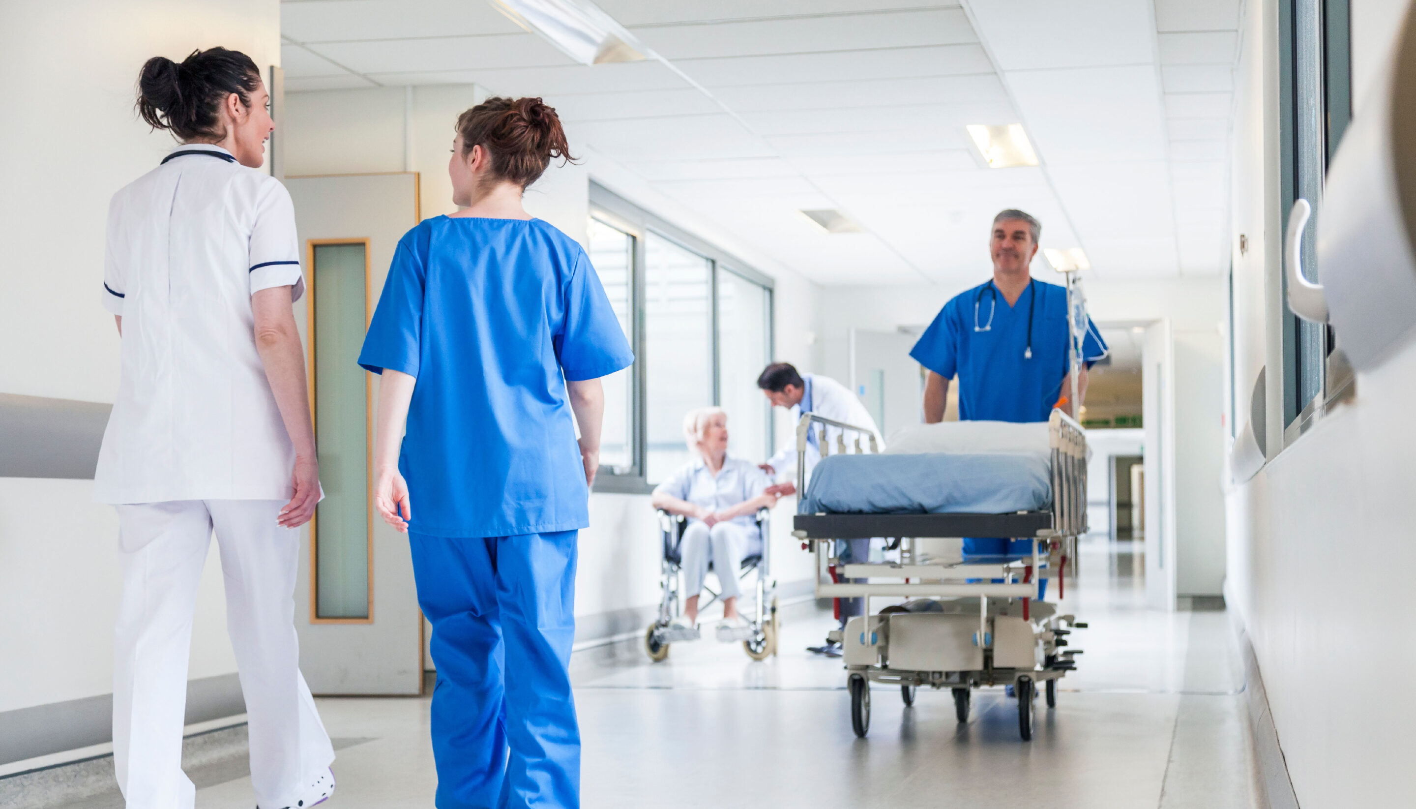 A bright hospital corridor with healthcare staff moving between patients; two medical professionals walk together in conversation while another team member pushes a hospital bed and a patient in a wheelchair is being assisted in the background.