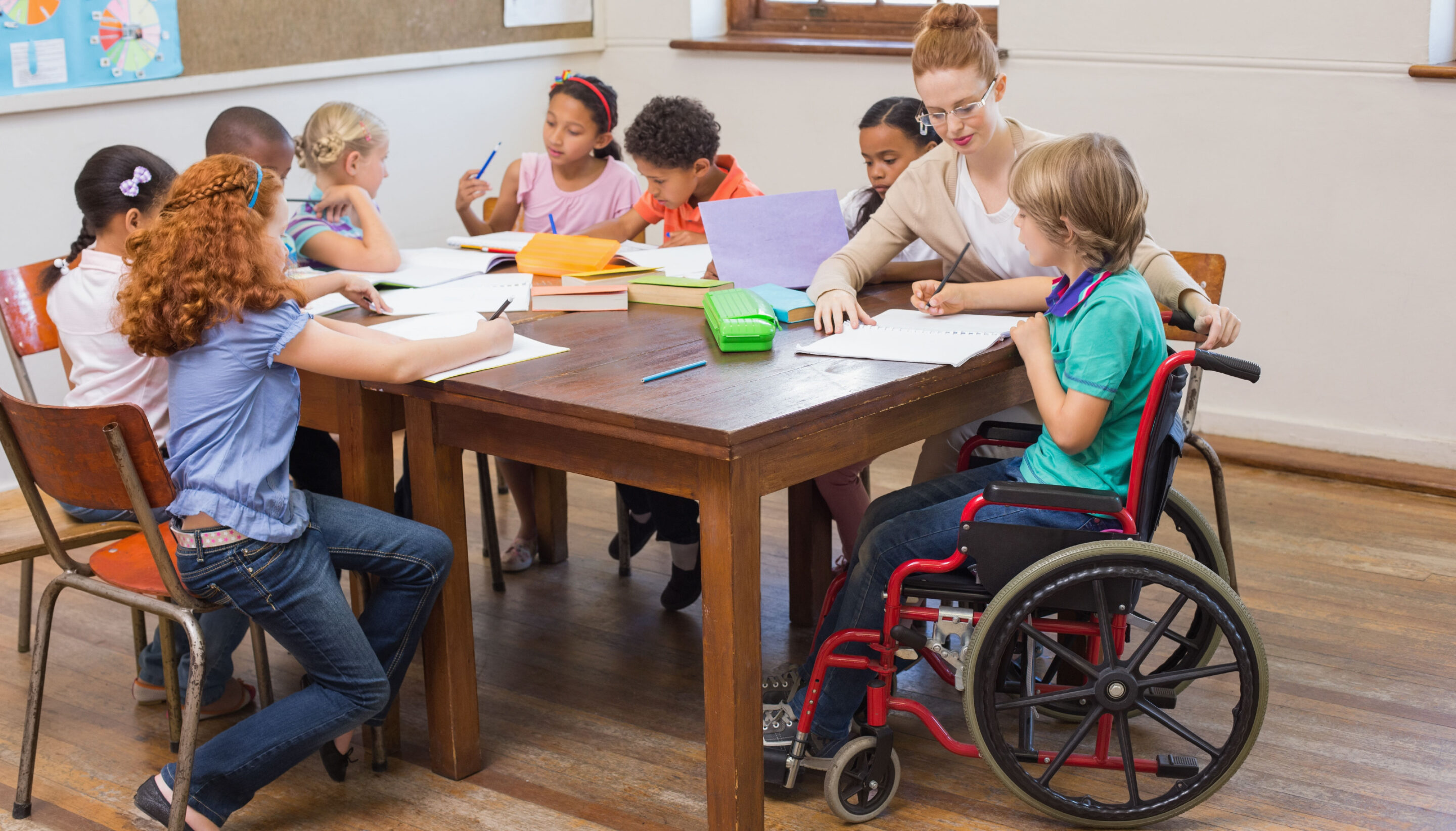 A group of young children sit around a large wooden table in a classroom, working on drawings and writing activities with a teacher demonstrating inclusion. Art supplies such as pencils and paper are spread across the table. One child is seated in a wheelchair, and the classroom has colourful posters and a corkboard on the wall.